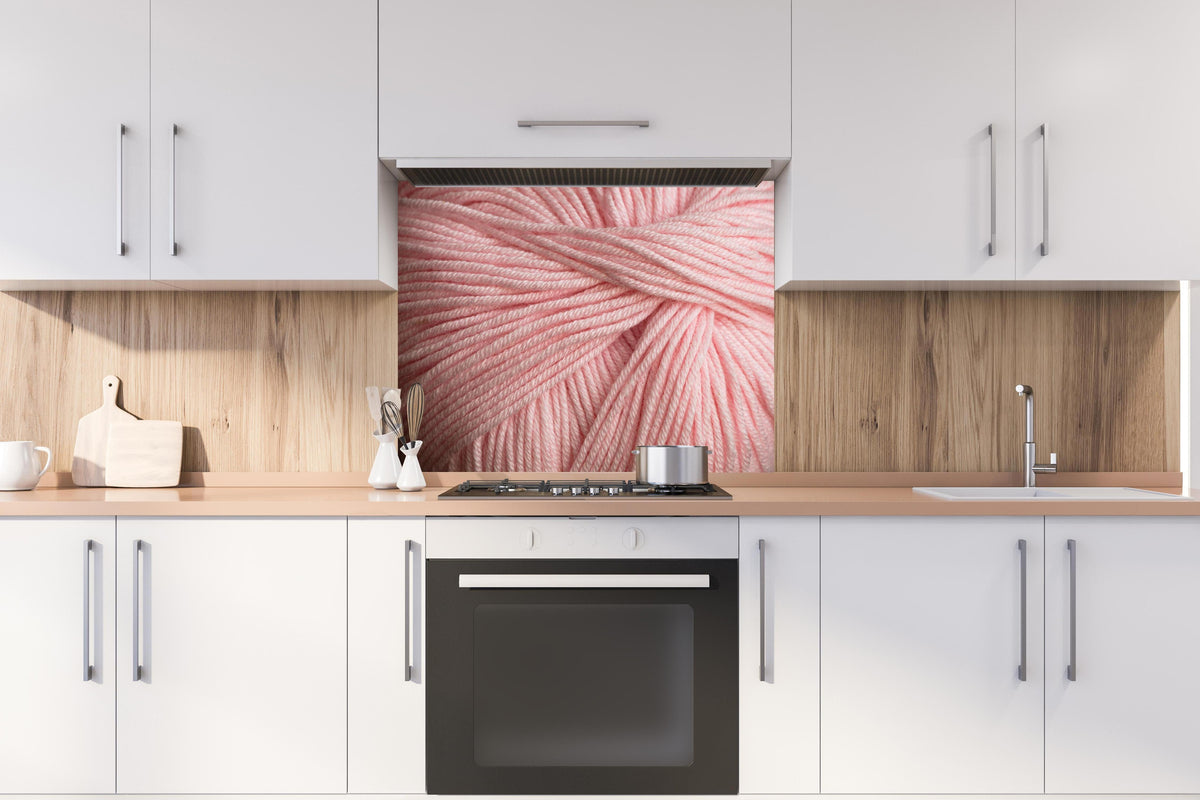 Spritzschutz - Nahaufnahme eines rosa Garnknäuels hinter einem Cerankochfeld zwischen Holz-Kochutensilien
