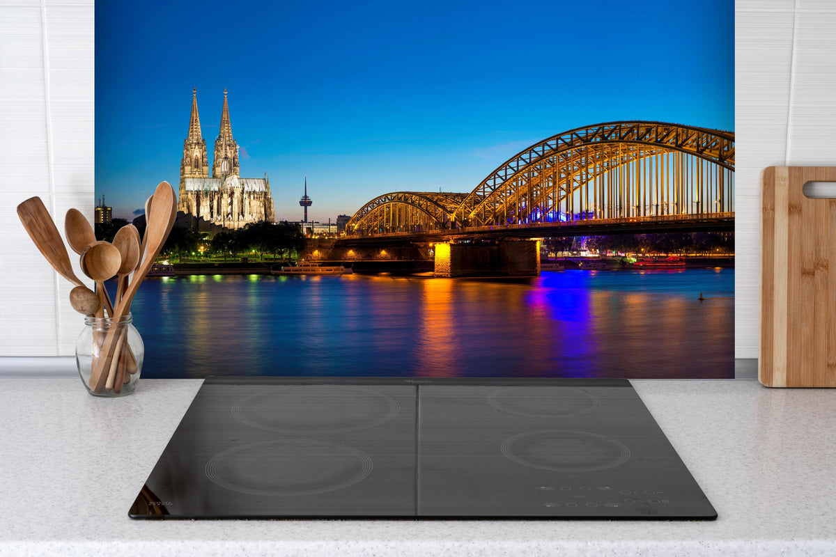Spritzschutz - Hohenzollern Brücke Köln in der Nacht hinter einem Cerankochfeld zwischen Holz-Kochutensilien
