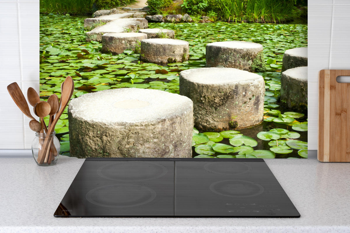 Spritzschutz - Stein-Zen-Pfad umhüllt von Wasserpflanzen hinter einem Cerankochfeld zwischen Holz-Kochutensilien
