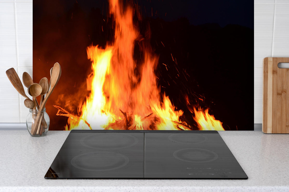 Spritzschutz - gewaltiges outdoor Lagerfeuer  hinter einem Cerankochfeld zwischen Holz-Kochutensilien
